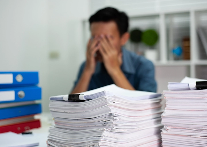 man with hands on face as if he has a headache while a stack of papers sits in front of him