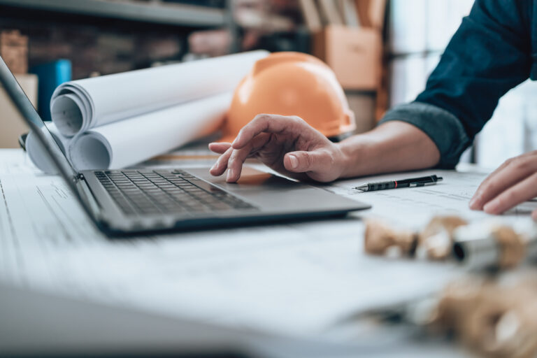 Close-up of hands typing on a laptop with rolled blueprints and an orange hard hat in the background.