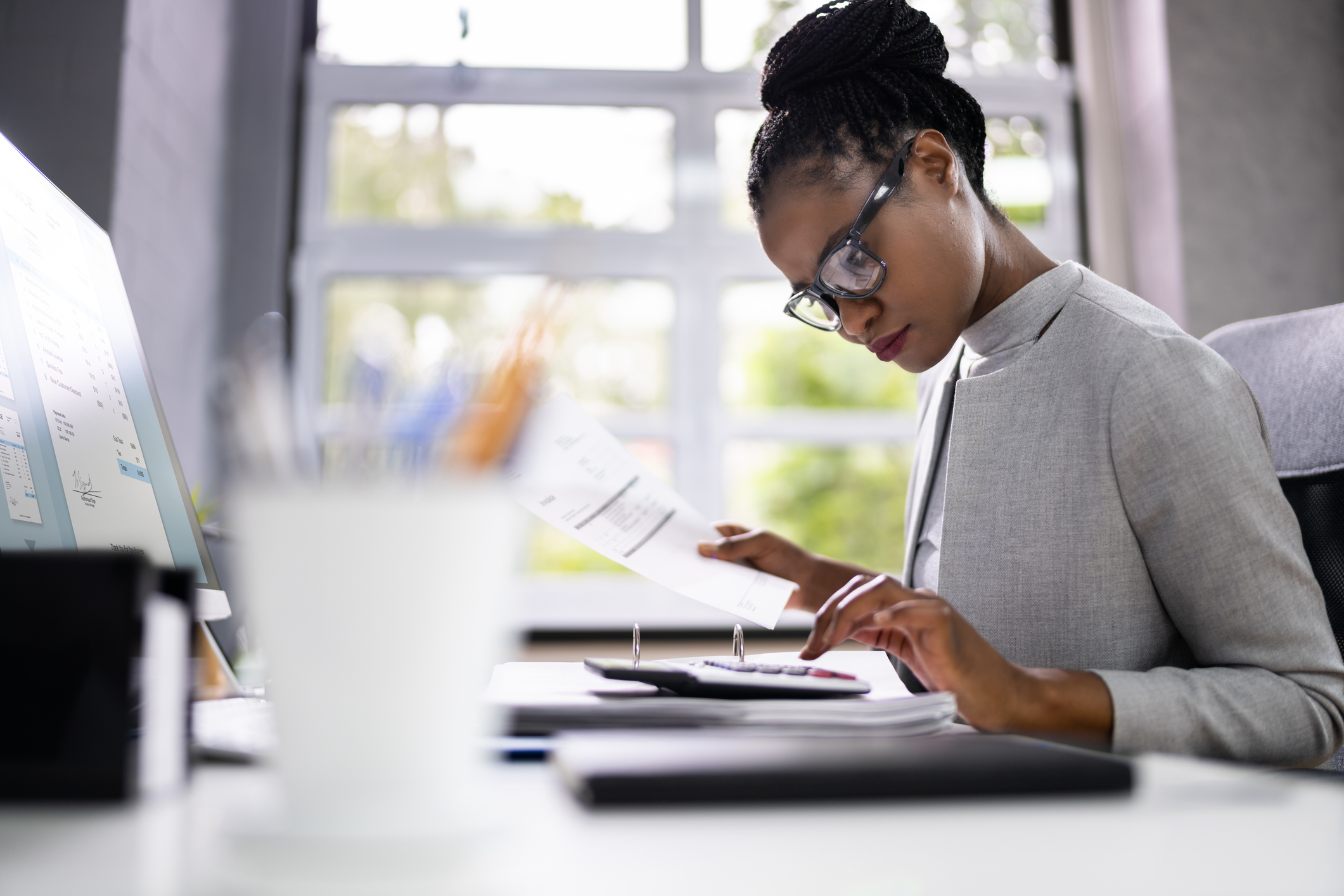 Professional woman in a gray blazer reviews documents at a desk, with a calculator nearby in a bright office.