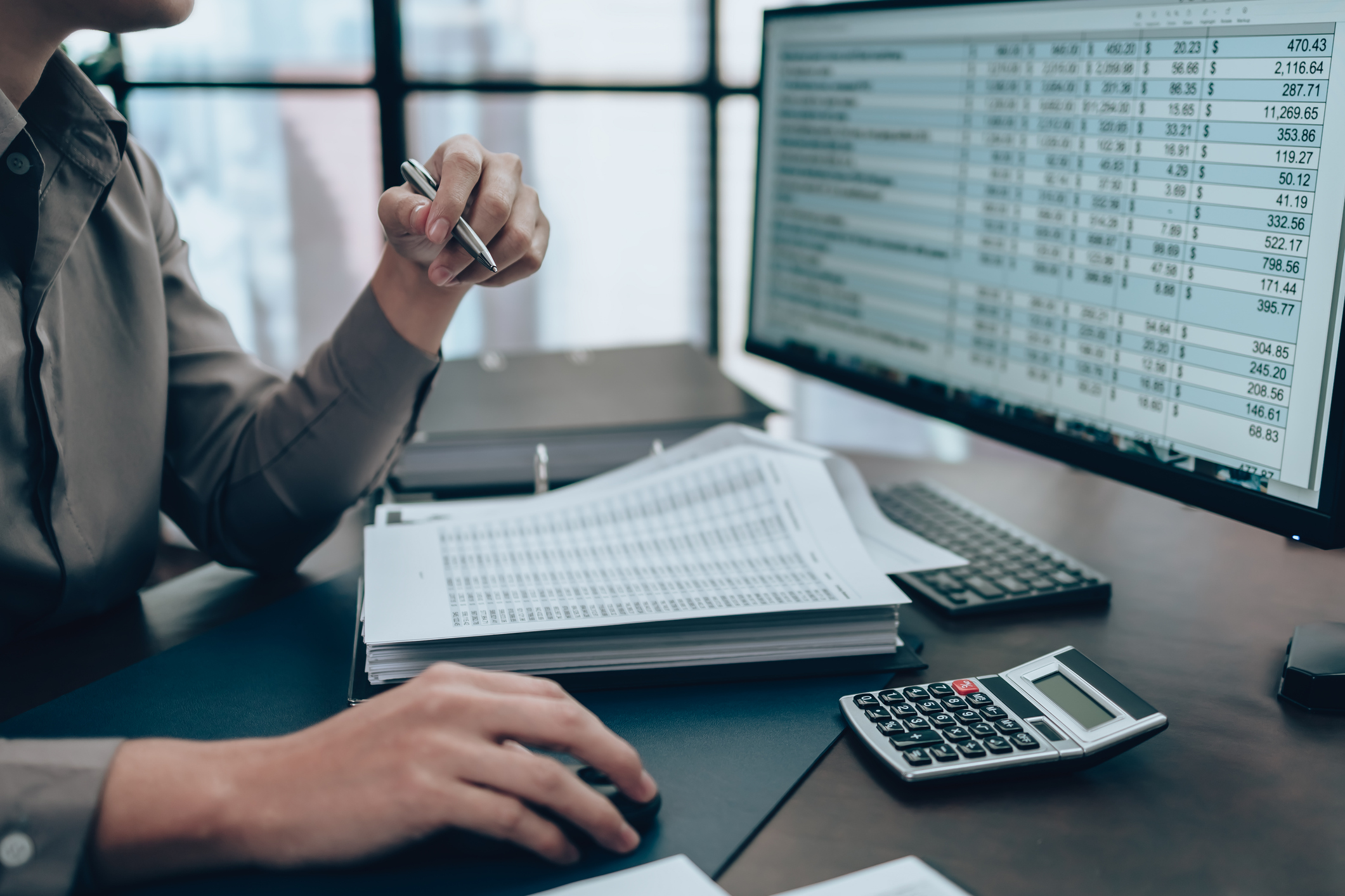 Person uses a pen while reviewing printed financial documents at a desk, with a calculator and a large spreadsheet on a monitor nearby.